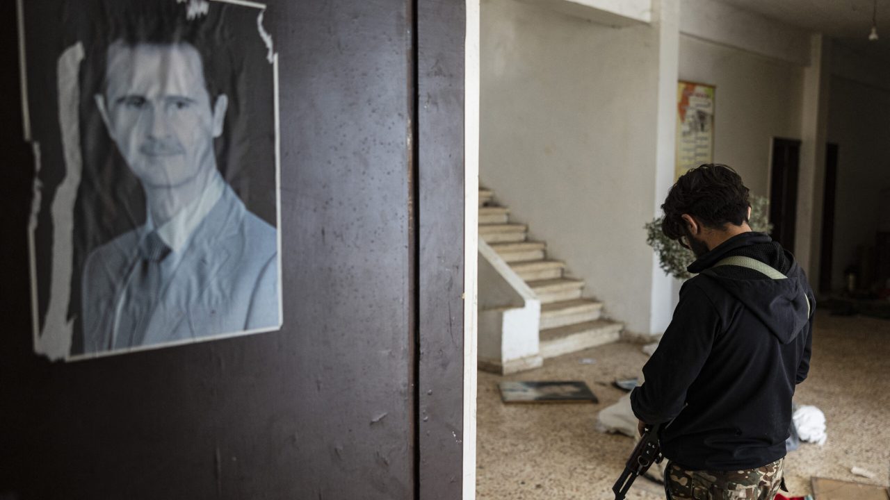 A rebel fighter walks past a portrait of toppled Syrian president Bashar al-Assad at the 4th Division barracks of the dismantled Syrian army, in the town of Dummar near Damascus, on December 23, 2024. Three rights group on December 23, appealed to Syria's new rulers to urgently preserve evidence of atrocities committed under former president Bashar al-Assad. (Photo by Sameer Al-DOUMY / AFP)