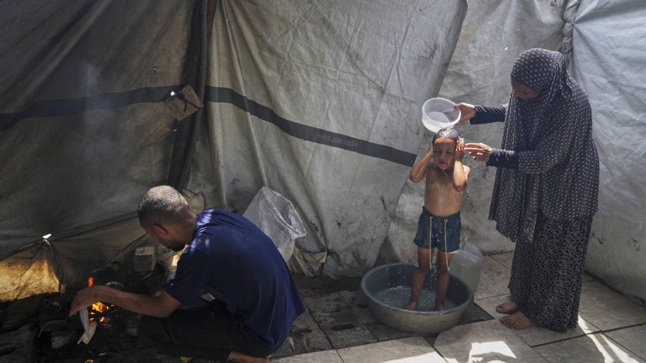 Enaam Al Majdoub uses water collected from a distribution point to bathe her 3-year-old granddaughter, Jourieh, while her son Zaki uses some of the water for cooking in their family tent in Gaza City on Tuesday, August 12, 2025. (AP Photo/Jehad Alshrafi)