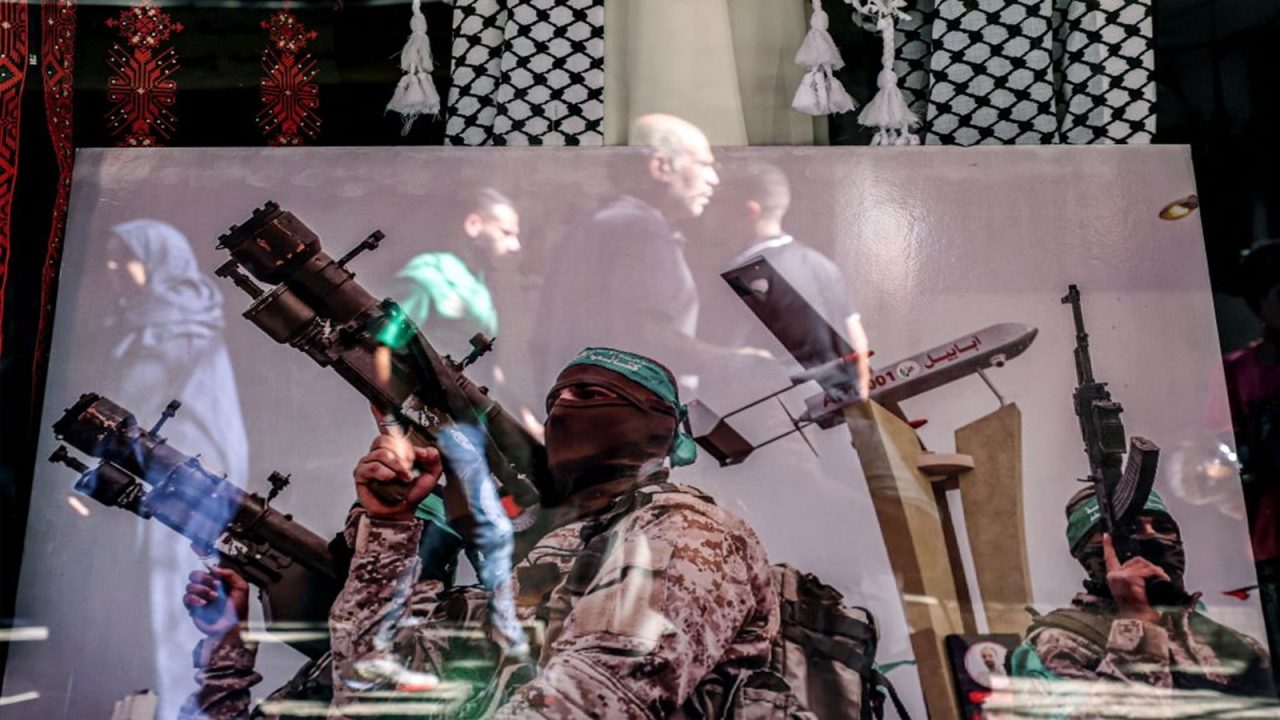 A reflection shows people walking past a poster in a shop window showing a fighter of the Izzedine al-Qassam Brigades, the military wing of the Palestinian Hamas movement, carrying a rocket launcher and posing near a mockup of a kamikaze drone along an alley at the Shatila camp for Palestinian refugees in the southern suburb of Beirut on November 7, 2023 amid ongoing battles between Israel and the Palestinian militant group Hamas in the Gaza Strip. (Photo by AHMAD AL-RUBAYE / AFP) (Photo by AHMAD AL-RUBAYE/AFP via Getty Images)
