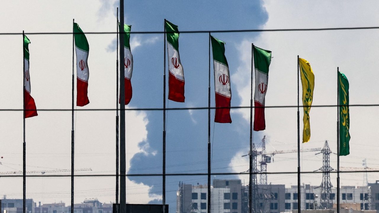 Flags flutter along a bridge as a plume of heavy smoke and fire rise from an oil refinery in southern Tehran, after it was hit in an overnight Israeli strike, on June 15, 2025. Iran fired a fresh barrage of missiles at Israel, state television announced early on June 15, as the rivals exchanged fire for a third day. (Photo by ATTA KENARE / AFP)