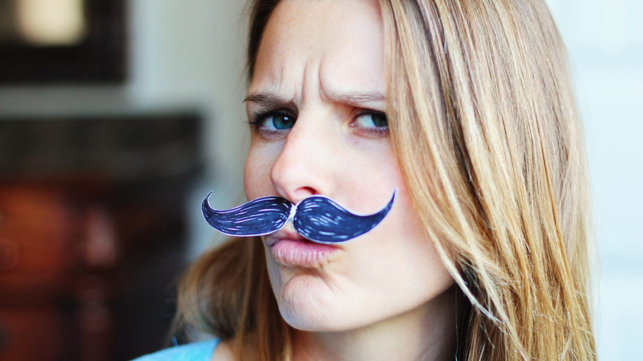 Close up of young woman with fake mustache under her nose. She is making silly face.